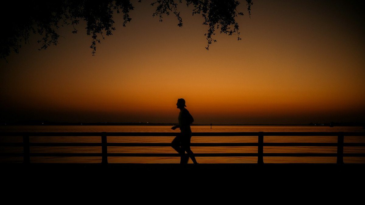 Silhouette sombre d'un coureur longeant une rambarde au bord de l'eau, sous un ciel de crépuscule