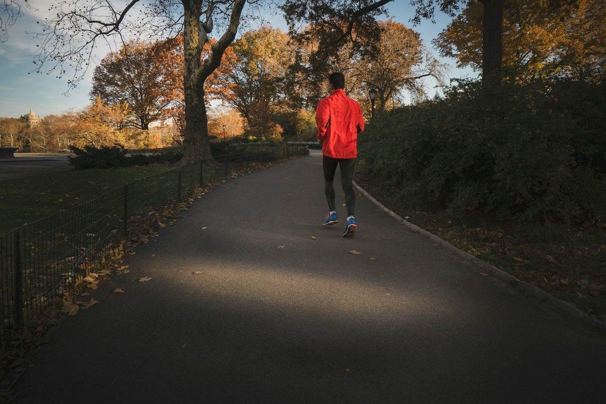 Coureur vu de dos en veste rouge sur une allée de parc bordée d'arbres d'automne
