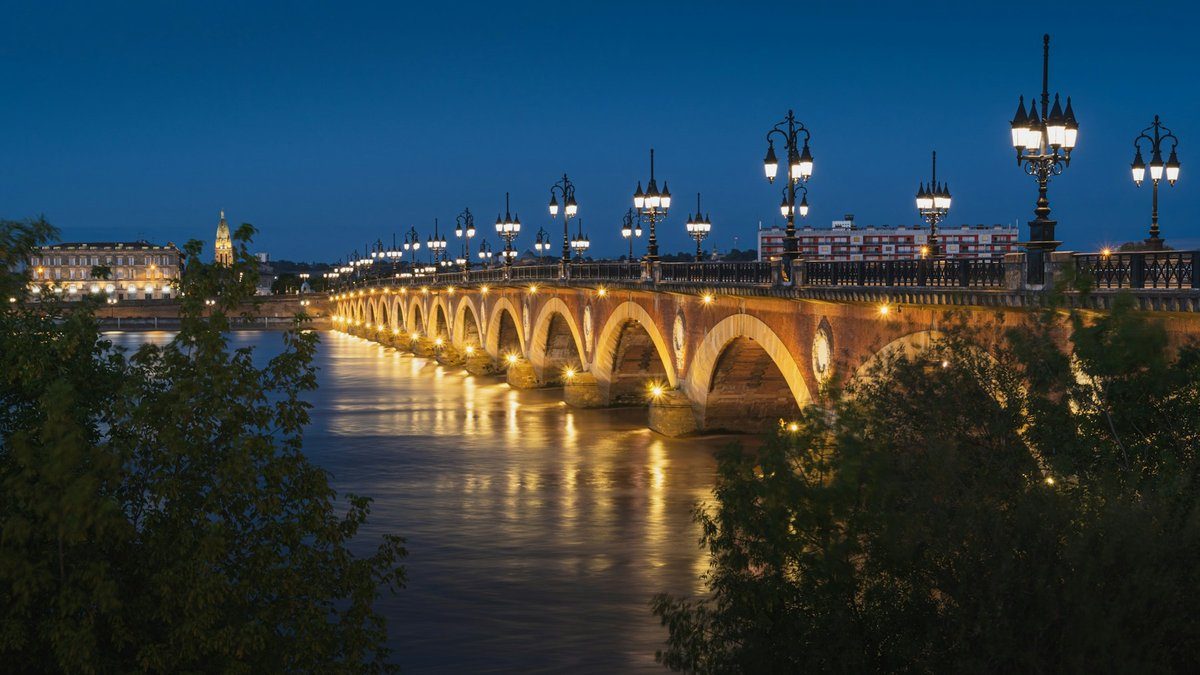 Le pont de pierre de Bordeaux illuminé, ses arches reflétées dans la Garonne sous un ciel bleu sombre